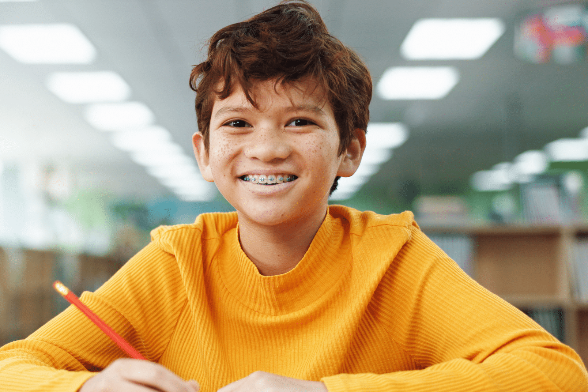 A smiling young boy with dental braces sits at a desk in a library,